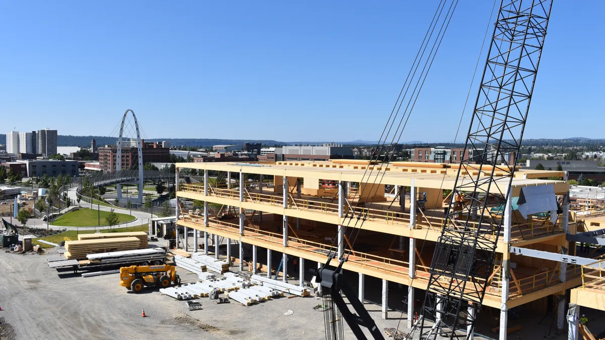 An aerial shot of an under-construction building. Wood is visible from a distance as the frame of the building is erected on a sunny day.