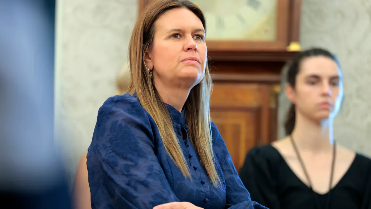 A person stands with arms crossed as another person looks on.