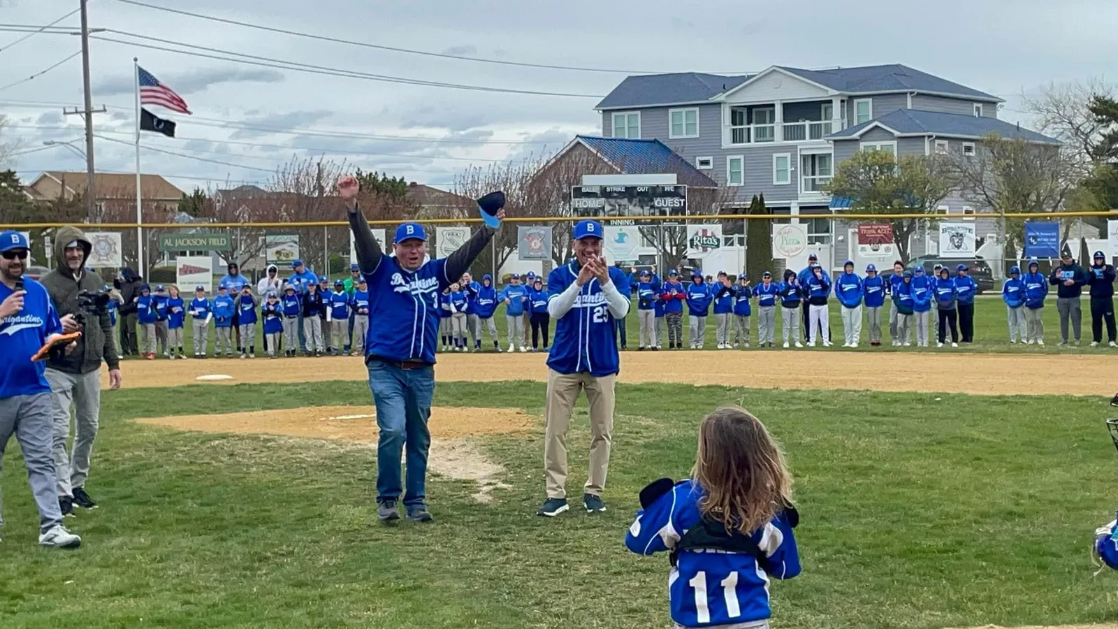 A man superintendent in a blue baseball jersey throws the first pitch at a youth recreational baseball game. Other adults and child players are shown around the baseball diamond in blue jerseys.