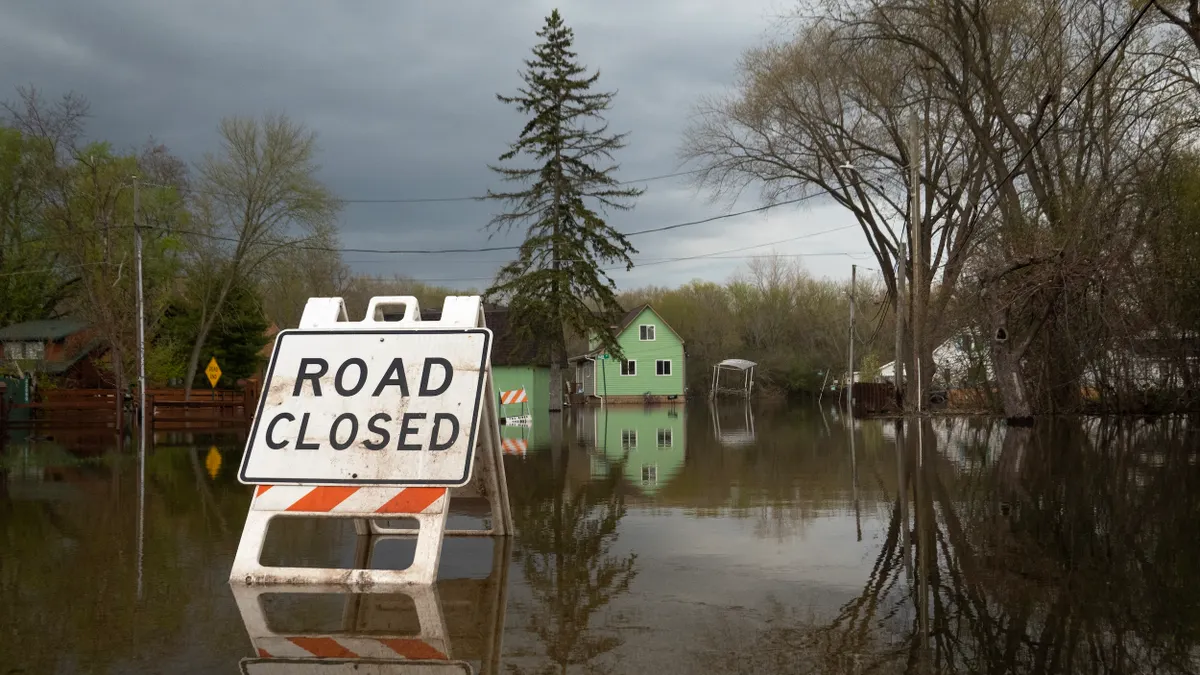A sign reading "road closed" on a flooded road with damaged homes behind.