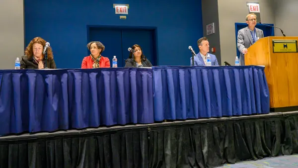Four people at a desk with microphones and a person standing at a podium.