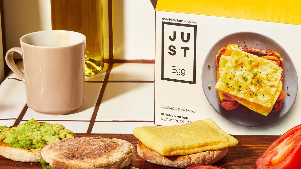 An English muffin breakfast sandwich with Just Egg folded patty, tomato and avocado and a split English muffin with a folded egg on one side and avocado on the other sit on a dark wood cutting board