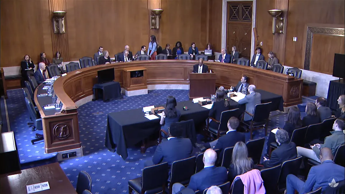 Overhead view of a senate committee room with several senators seated at a rounded dais and three witnesses at a table facing them with a seated audience behind.