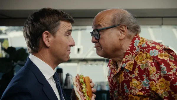 Danny DeVito and Eli Manning stand face-to-face as Manning holds a sub sandwich.