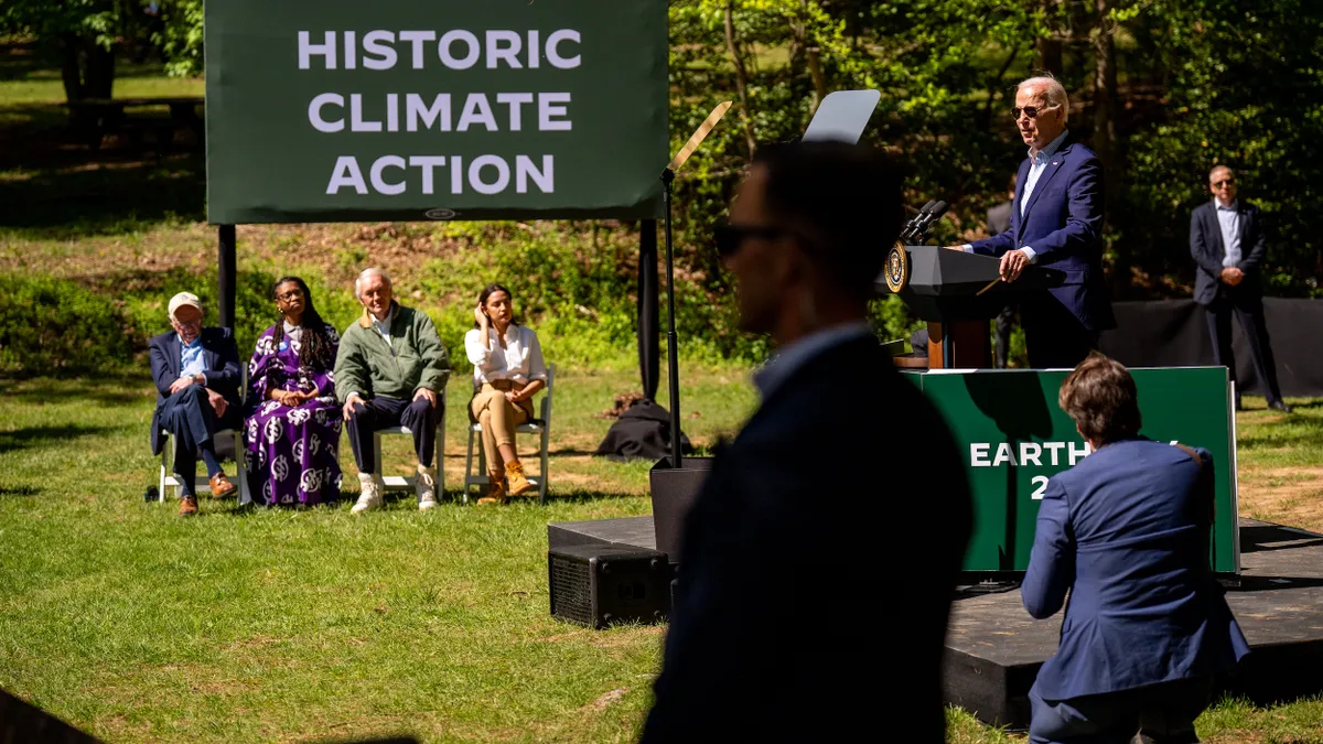 Four people, including members of Congress, sit in chairs under a sign that reads "President Joe Biden Historic Climate Action." Biden stands on a small stage to the right.