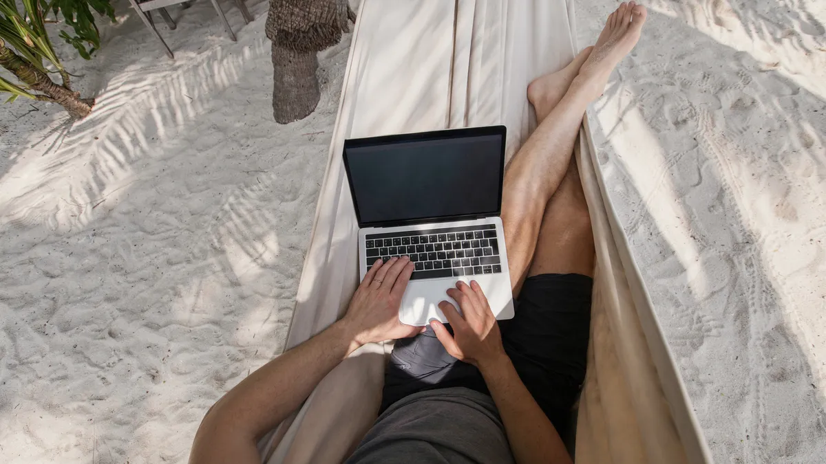 An individual works on a laptop while relaxing in a hammock on the beach.