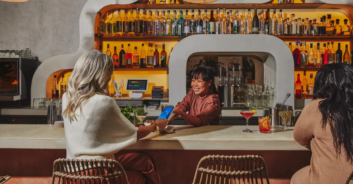 Two people are seated at a bar. A bartender uses a mobile device to scan a one of the customer&#x27;s credit card while smiling.