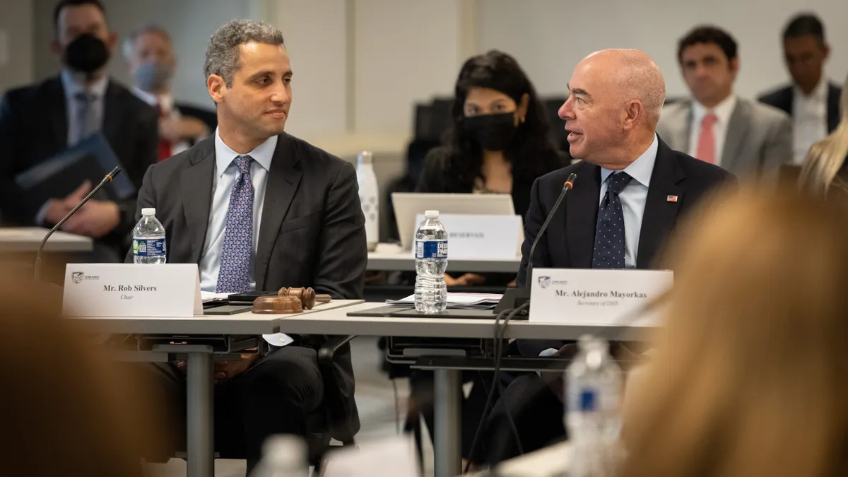 Rows of people sit at tables with name cards in front of each of them. The two people in the foreground look at each other as they converse.