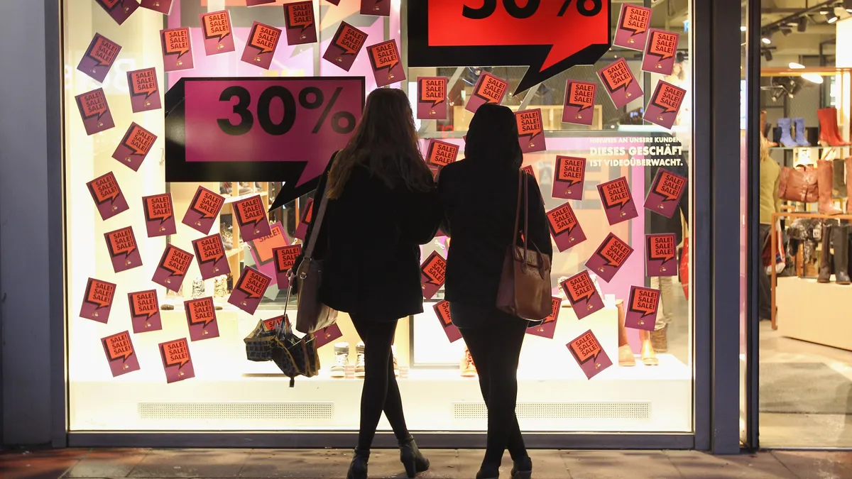 Shoppers stand in front of a store window with large signs boasting sales