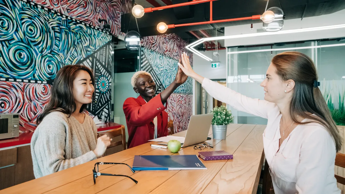 Three co-workers sit at a conference table; two people exchange a high-five