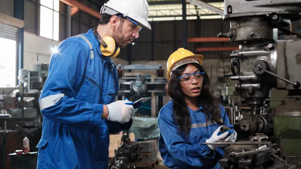 Two industrial workers in protective and safety uniforms and hardhats working with metalwork machines in a manufacturing factory.