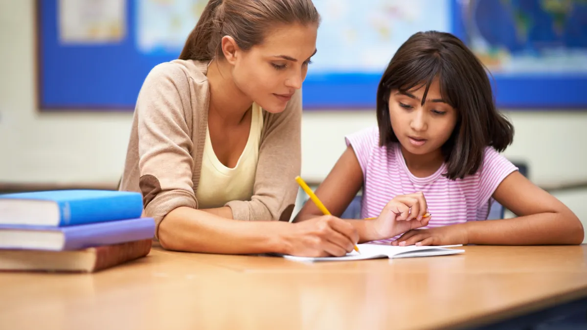 A teacher works with a student on an assignment while sitting together at a desk in a classroom.