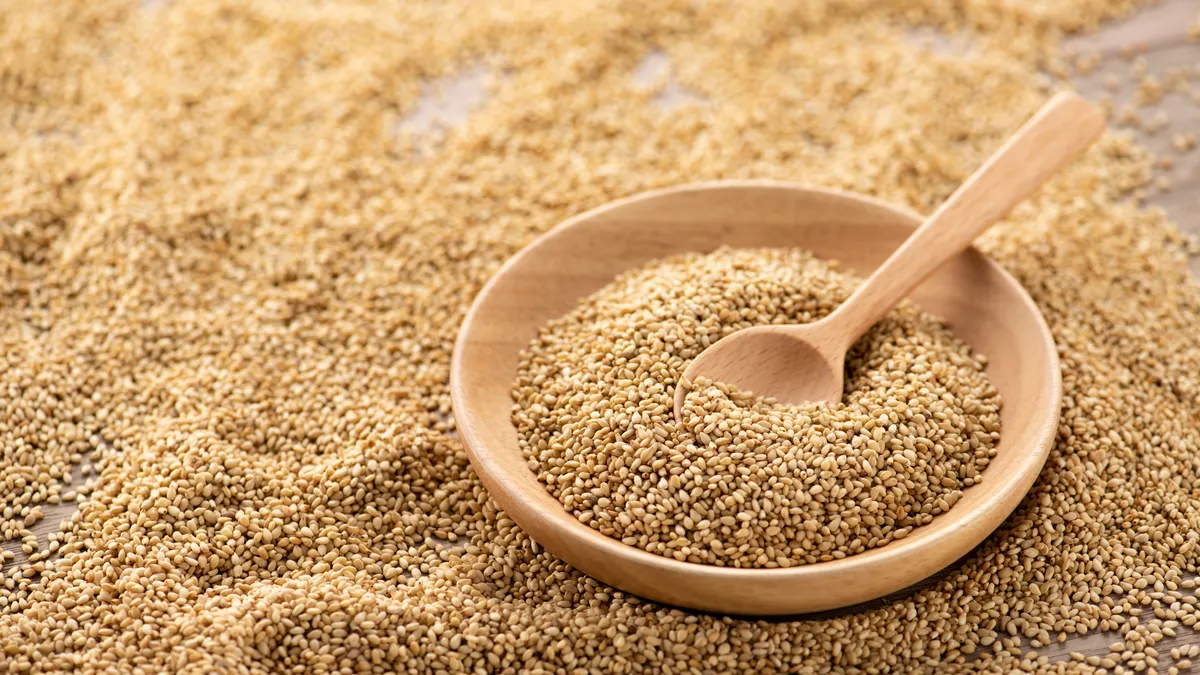 Sesame seeds cover a wooden table. A lighter wood bowl full of sesame seeds with a matching wooden spoon inside sits among them.