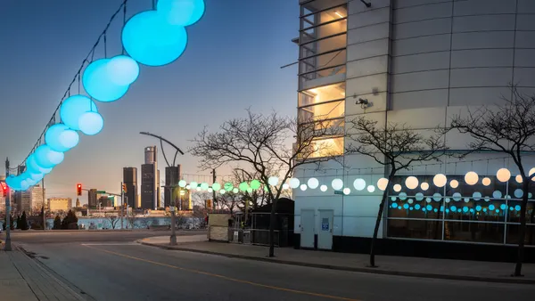 Strings of blue, orange and green hanging lanterns over a street with tall buildings in the background