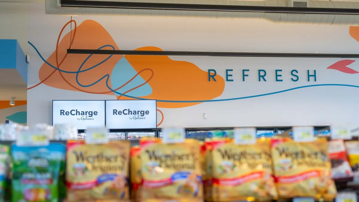 A photo of the wall inside of a store, seen over the top of several shelving units. Signs on the wall say "Refresh" and "Recharge by Gelson's."