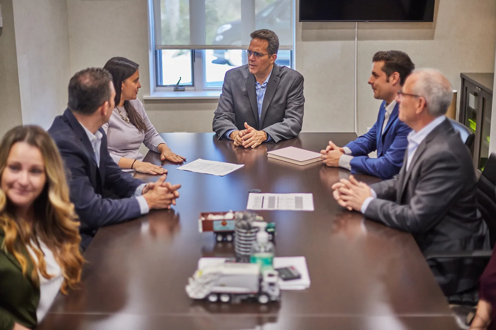 A group of people in suits and formal attire sitting around a conference table, trash truck models in the center