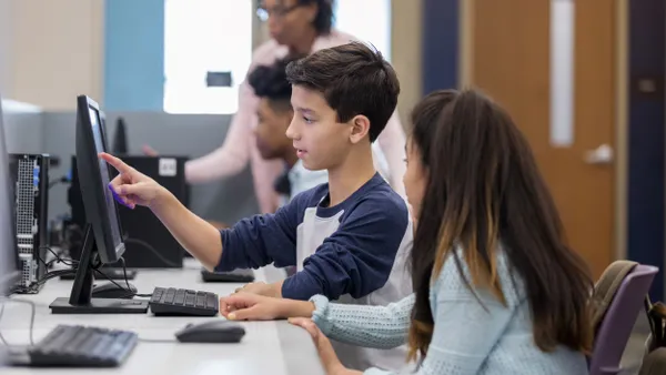 A middle school student points at a computer screen while a classmate looks over.