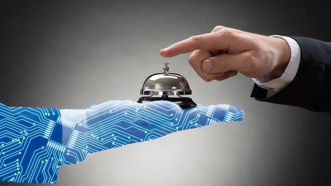 An extended robot arm holds a front desk bell while an extended human arm prepares to press down on the top