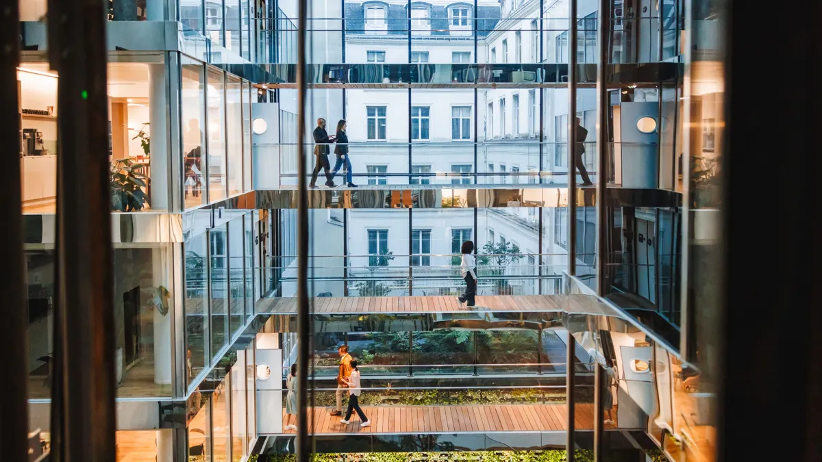 Men and women of varied ethnicities engage in professional tasks in a well-lit modern office, showcasing teamwork and individual focus, with urban views and greenery enhancing the workspace.