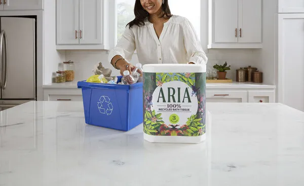 A person stands behind a kitchen counter where a package of Aria toilet paper has been placed next to a blue recycling bin.