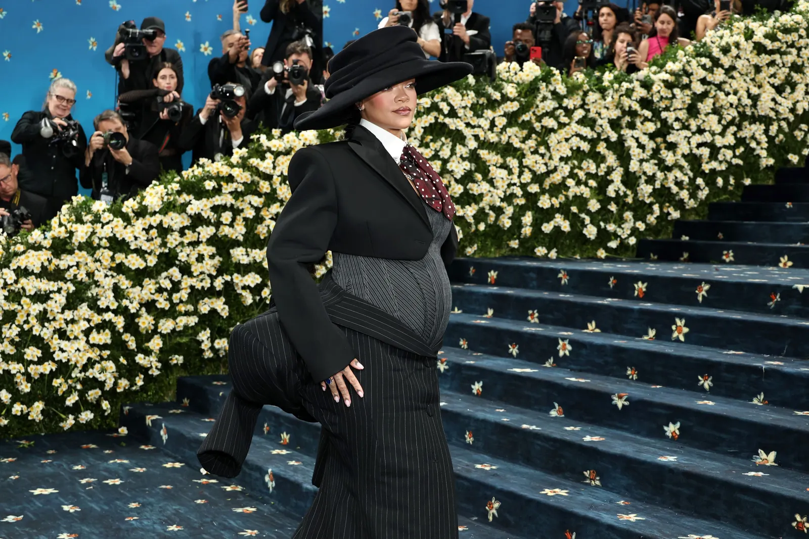 A woman in a wide-brimmed hat and pinstriped-maternity suit dress poses on a blue carpet.