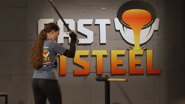 The back of a person with long hair in a gray t-shirt and black jeans, raising a thin sword above their head.