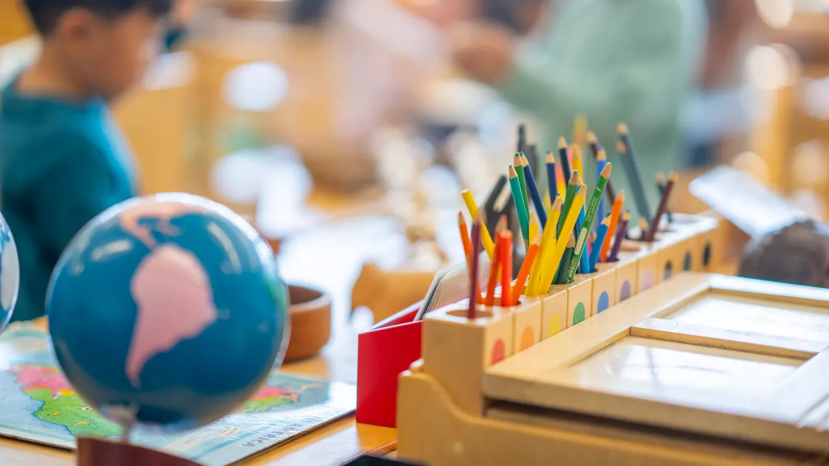 A photo of a elementary-level classroom. In the forefront is a desk with a globe and holder for colored pencils. In the background are young students but their images are blurred.