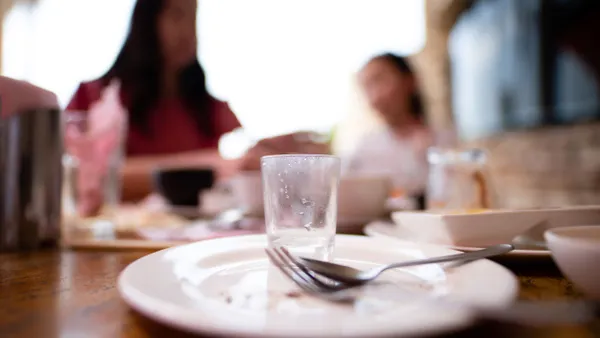 An empty plate is in focus at a table. In the blurred background, an adult and child are sitting at the same table.