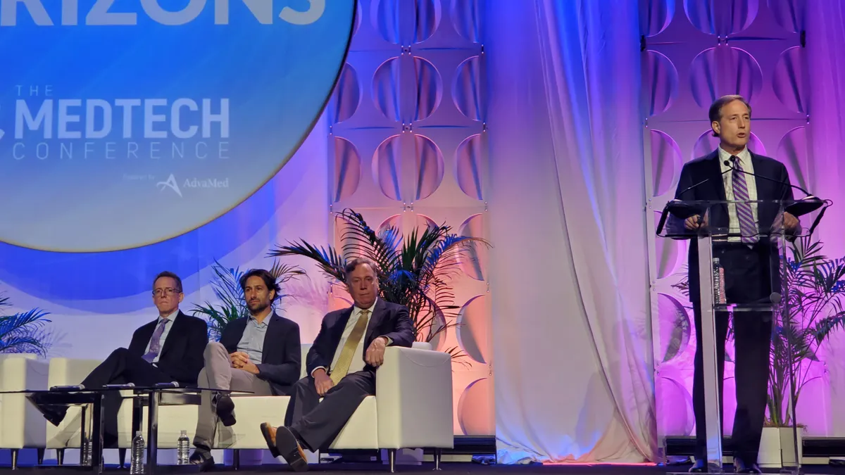 Three people sit on couches on a stage and one person speaks at a podium.