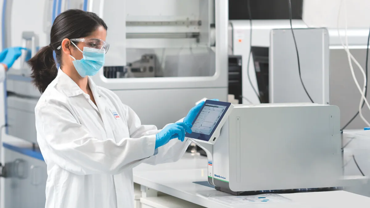 A person wearing goggles and a mask taps a tablet screen attached to a square machine.