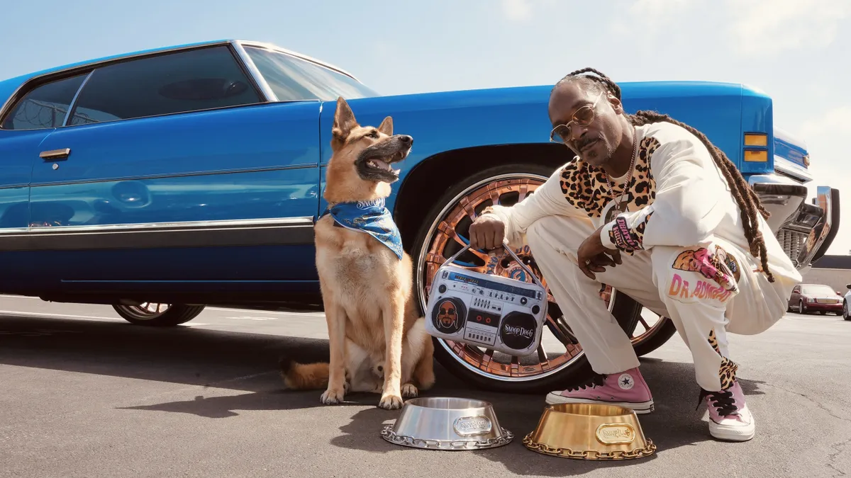 Snoop Dogg crouches down next to a car, a dog, and a gold and silver dog bowl looking like a baller.