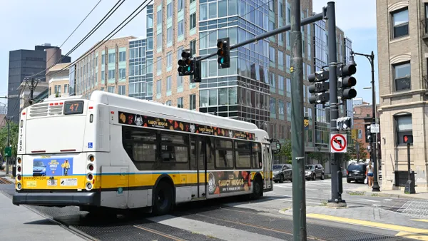 A white bus with a blue and yellow stripe seen from the rear as it drives through an intersection.