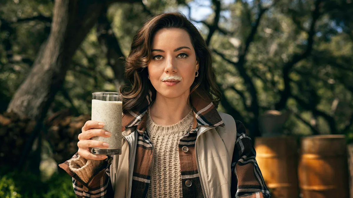 Aubrey Plaza sits in a forest, holding a glass of a thick white drink with black speckles in her right hand. On her upper lip, she has a "milk mustache" from this beverage.