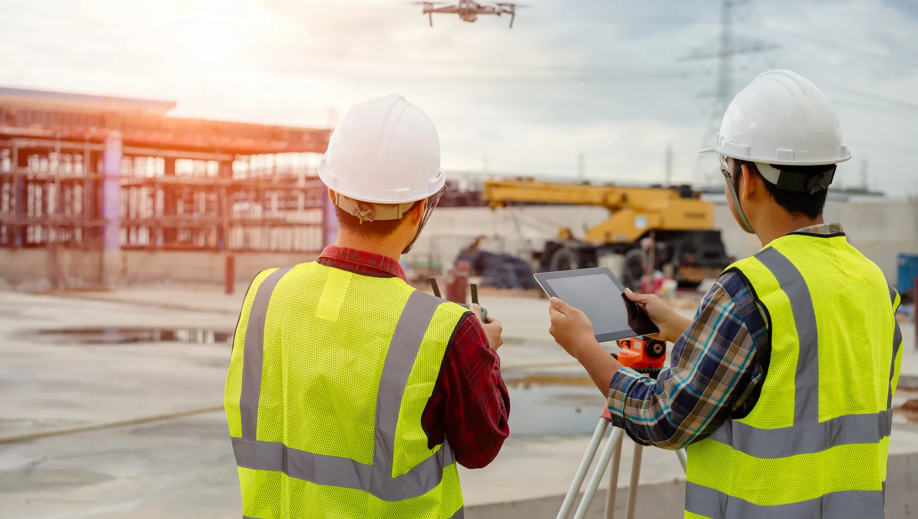 Two construction workers stand as one operates a drone. The drone is in the foreground of the picture, with the two workers who wear hardhats and safety vests.