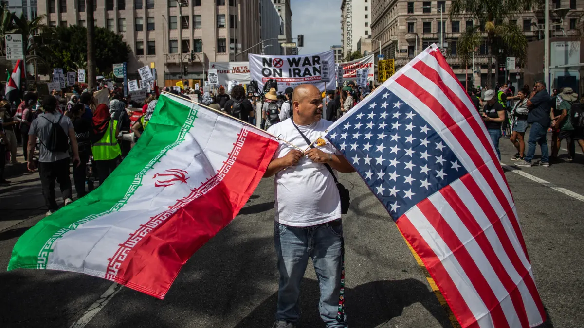 A man holds an upside-down American flag and an Iranian flag as he marches down a street. Behind him, other people hold signs, one of which says, "No War On Iran."