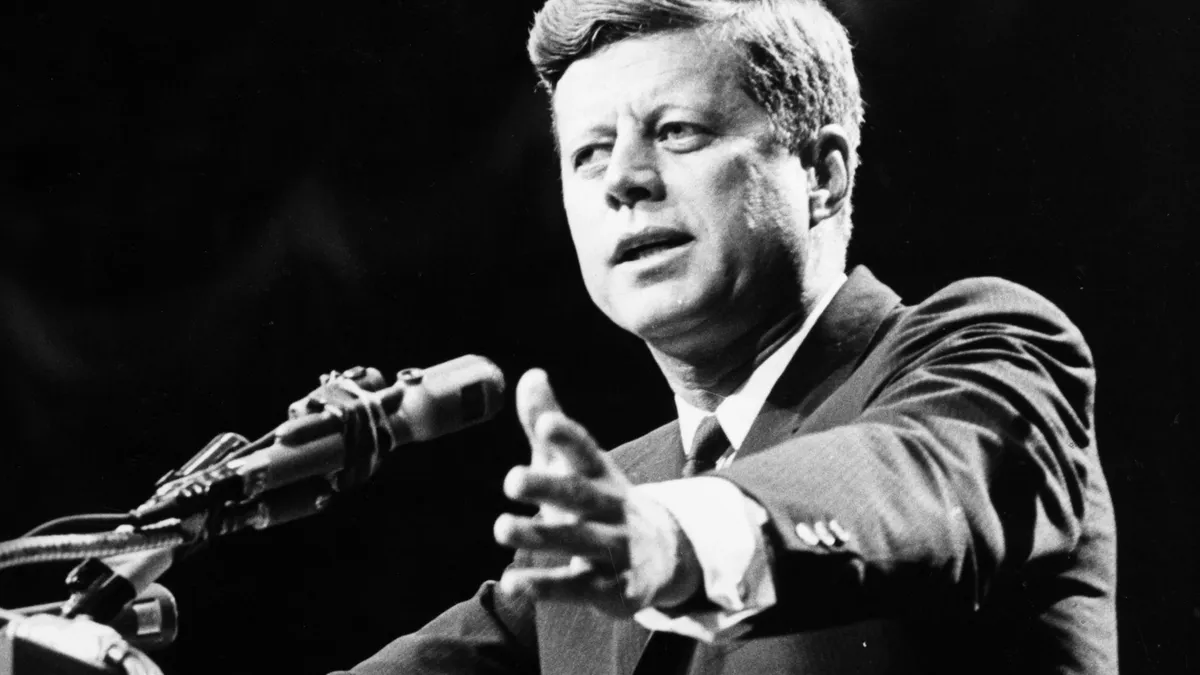 President John F. Kennedy is shown delivering an address at a podium in black and white.