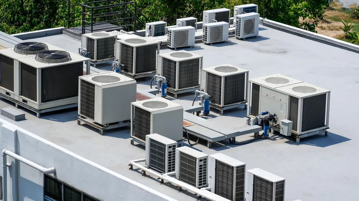 A flat rooftop is covered with various large air conditioning and ventilation units, arranged in neat rows with metal ducting, against a backdrop of green trees.