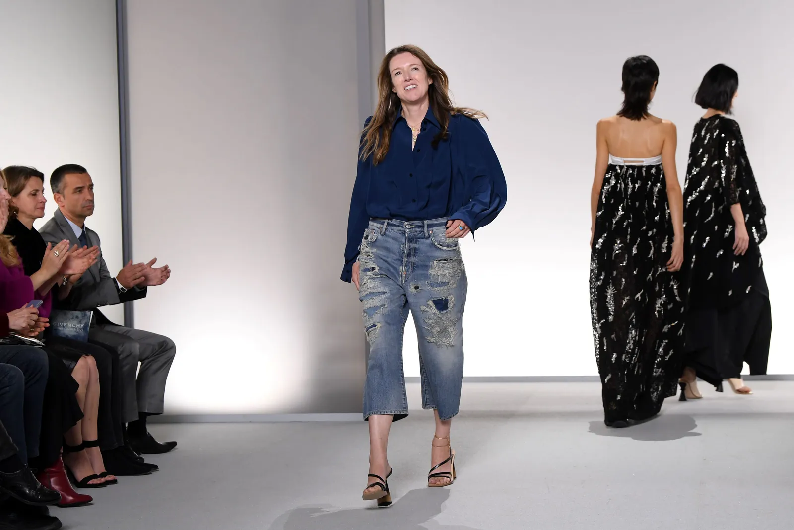 A woman in jeans walks down a runway smiling.