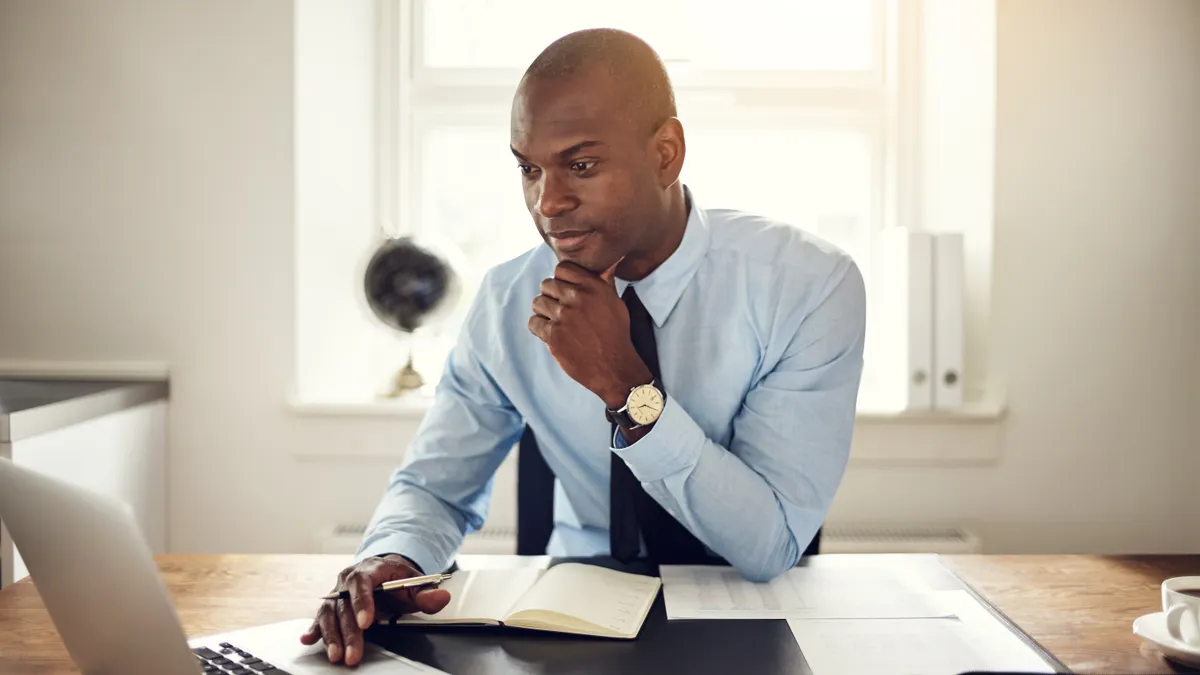 A Black business executive wearing a shirt and tie sitting at his desk in an office working online with a laptop