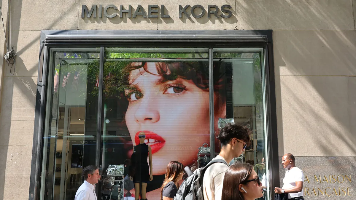 People walk past a Michael Kors store on a busy sidewalk during the day.