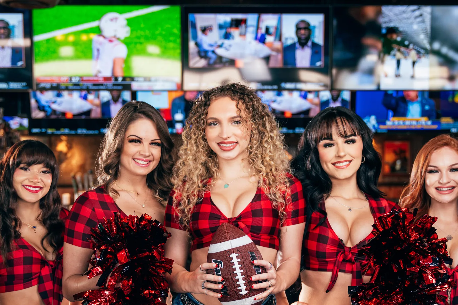 A photograph of several waitresses in plaid shirts in front TVs hanging from the sky.