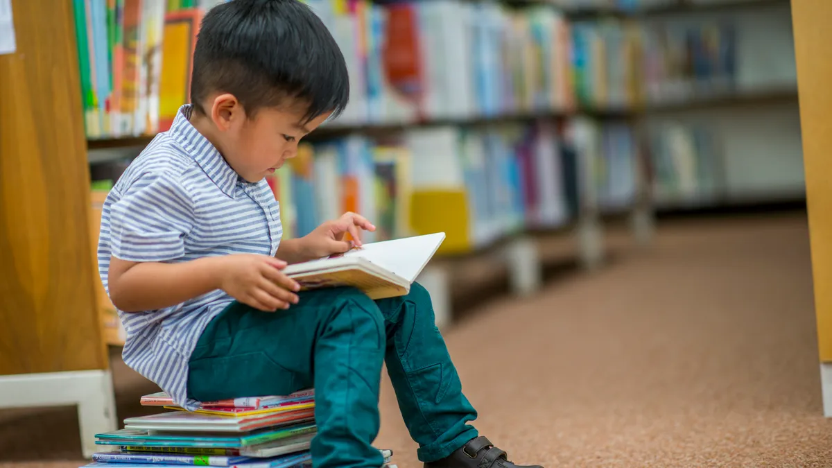 A young child sits on a stack of books near shelves of books. Child has a book on lap and is looking at book.