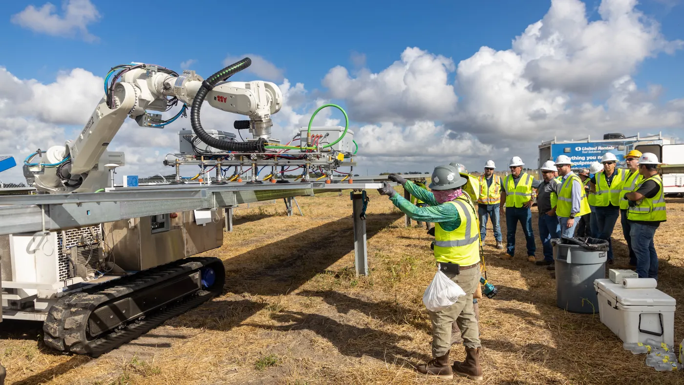 A large robot with a mechanical arm hefts a heavy solar panel while people in safety equipment watch.