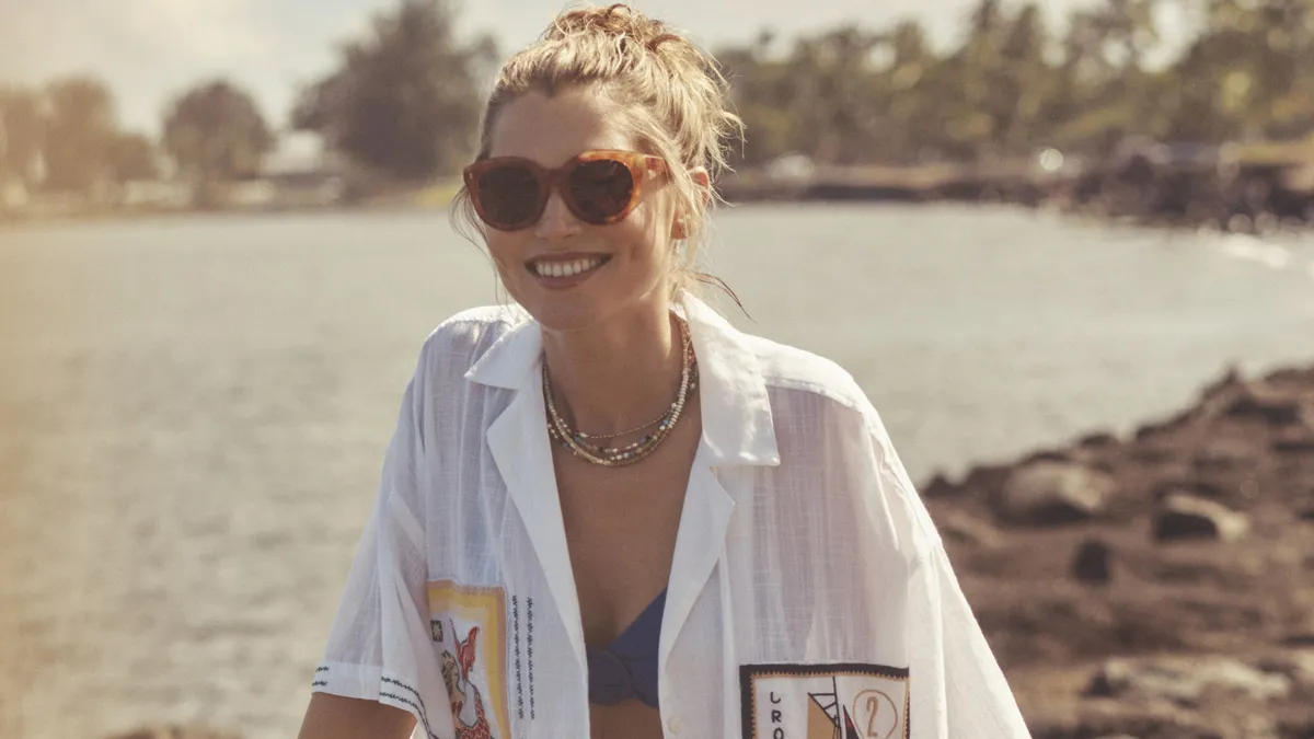 A model wears clothing from Antrhopologie's new resortwear line, including sunglasses, a blue bikini and an oversized button down featuring stamp graphics. The model rides a bike.