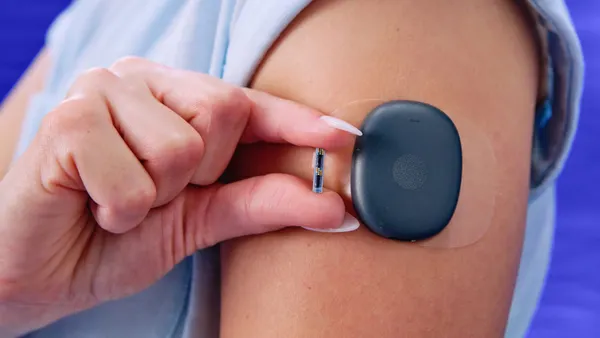 A person wears a round, black patch stuck to their upper arm. They hold a tiny cylinder between their thumb and pointer finger.