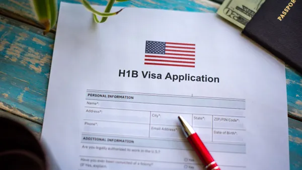 A printout application for an H-1B visa is on a table next to a pen and a passport with some cash sticking out.
