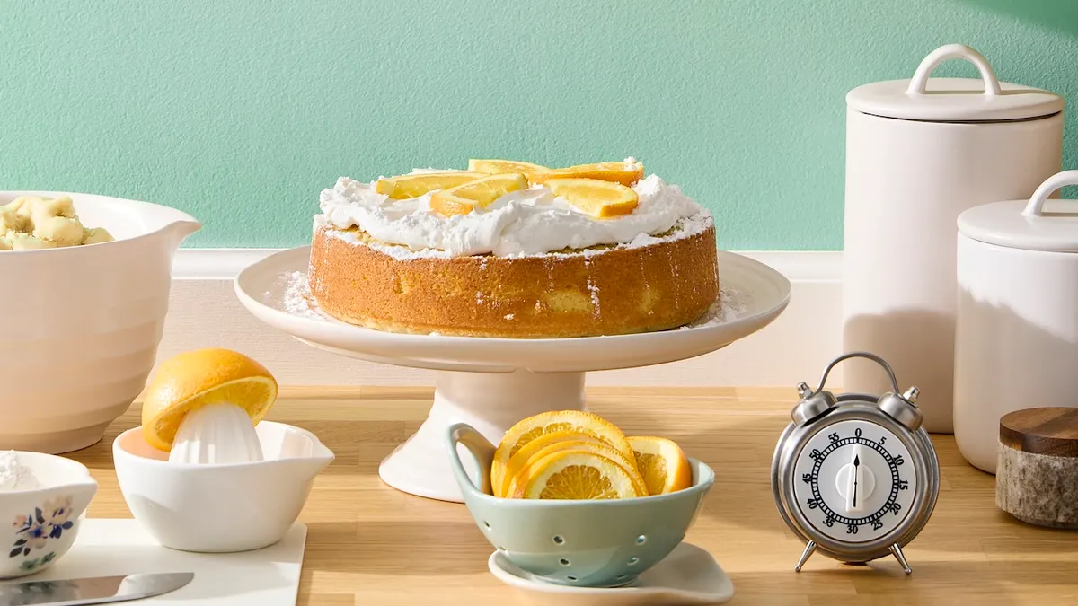 A cake on a white cake stand against a mint green wall. A bowl of lemon slices is positioned in front.