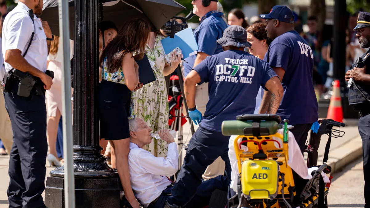 People crowd around a person sitting on the ground drinking water out of a plastic bottle. One person holds an umbrella over the person on the ground.