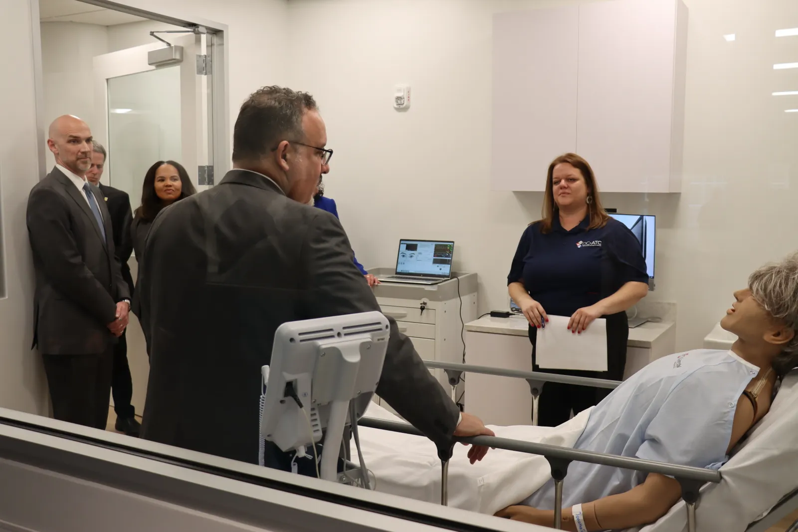 Adults are standing in a room with a glass window. A mannequin is laying in a bed in the room.