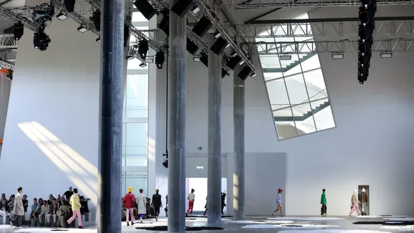 A line of models walk during the day in a cavernous room flanked by tall pillars.
